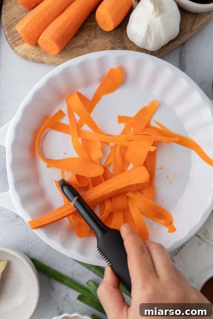 A vegetable peeler creating thin carrot ribbons from a fresh carrot over a bowl of water.