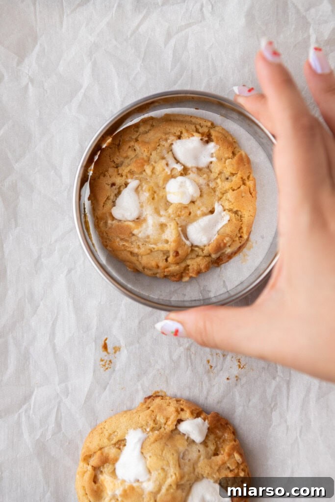 A hand using a cookie cutter to swirl a hot, freshly baked cookie into a perfect circle, containing melted marshmallow.