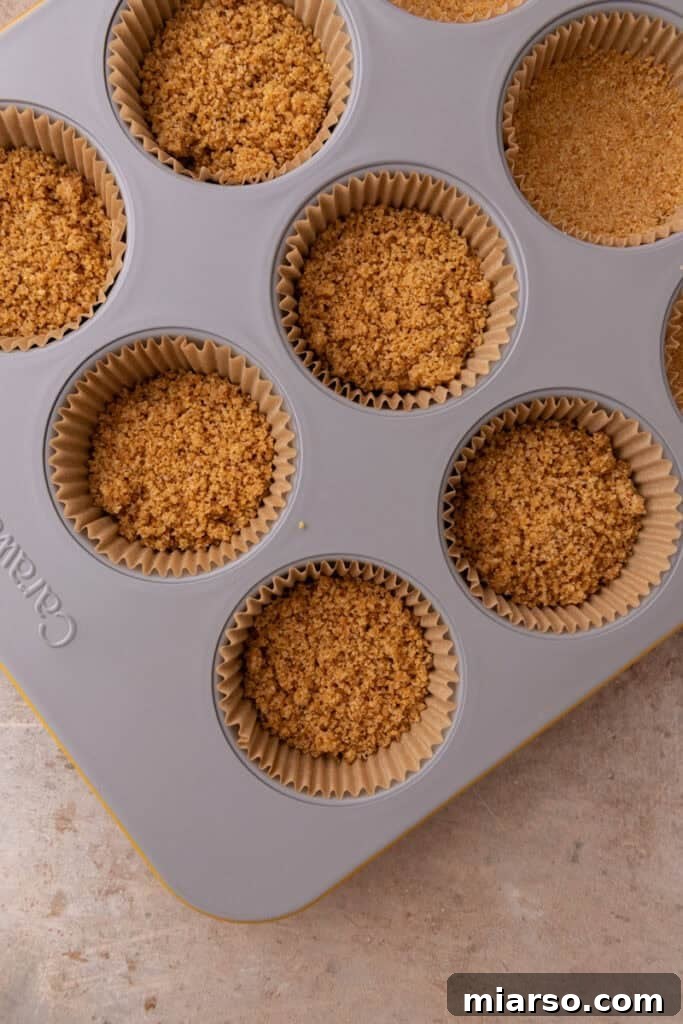 The graham cracker mixture is spooned into muffin liners, forming the base for the cheesecakes.