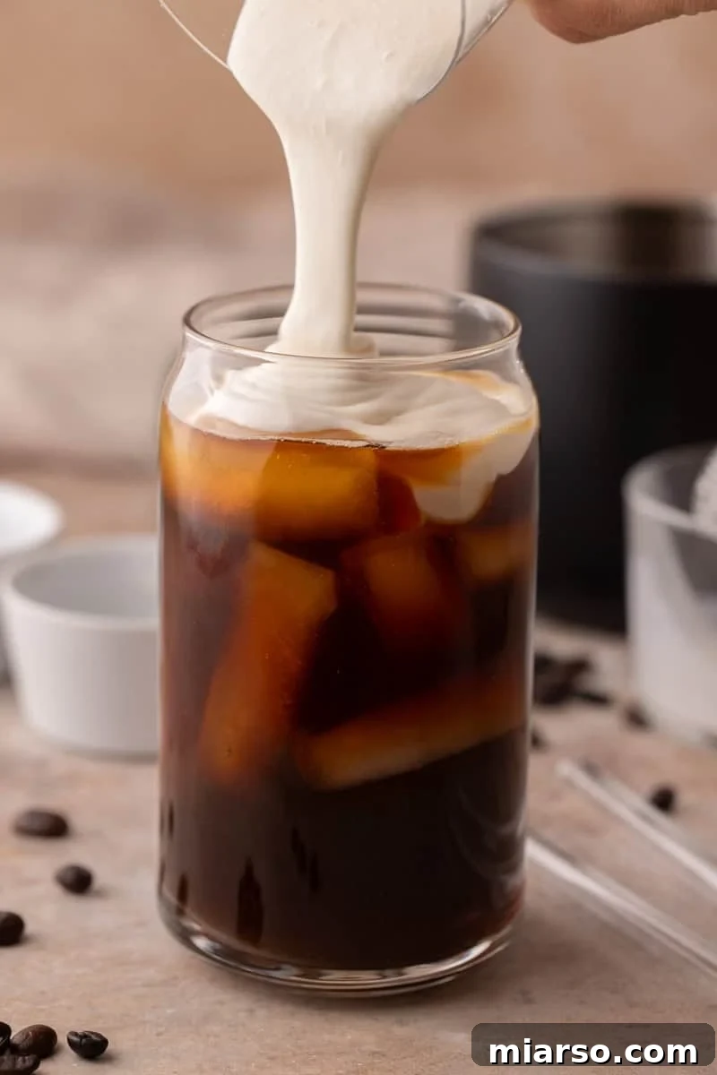 Starbucks Inspired High Protein Cold Foam 6 Thick, perfectly frothed homemade protein cold foam being poured gracefully over a glass of iced coffee, creating a visually appealing layer.