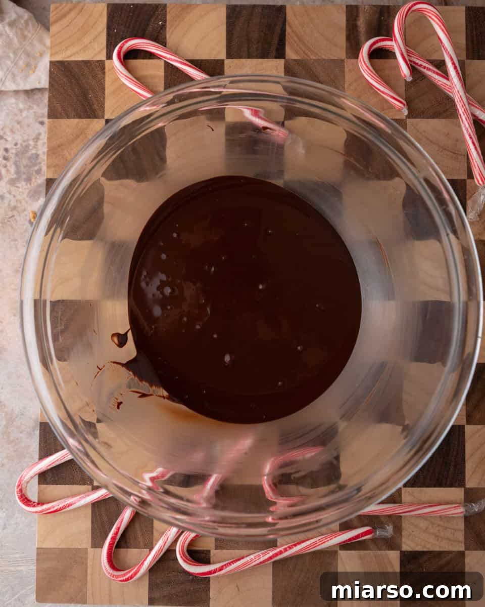 Smooth melted dark chocolate in a glass bowl on a wooden board decorated with candy canes, ready for the next step.