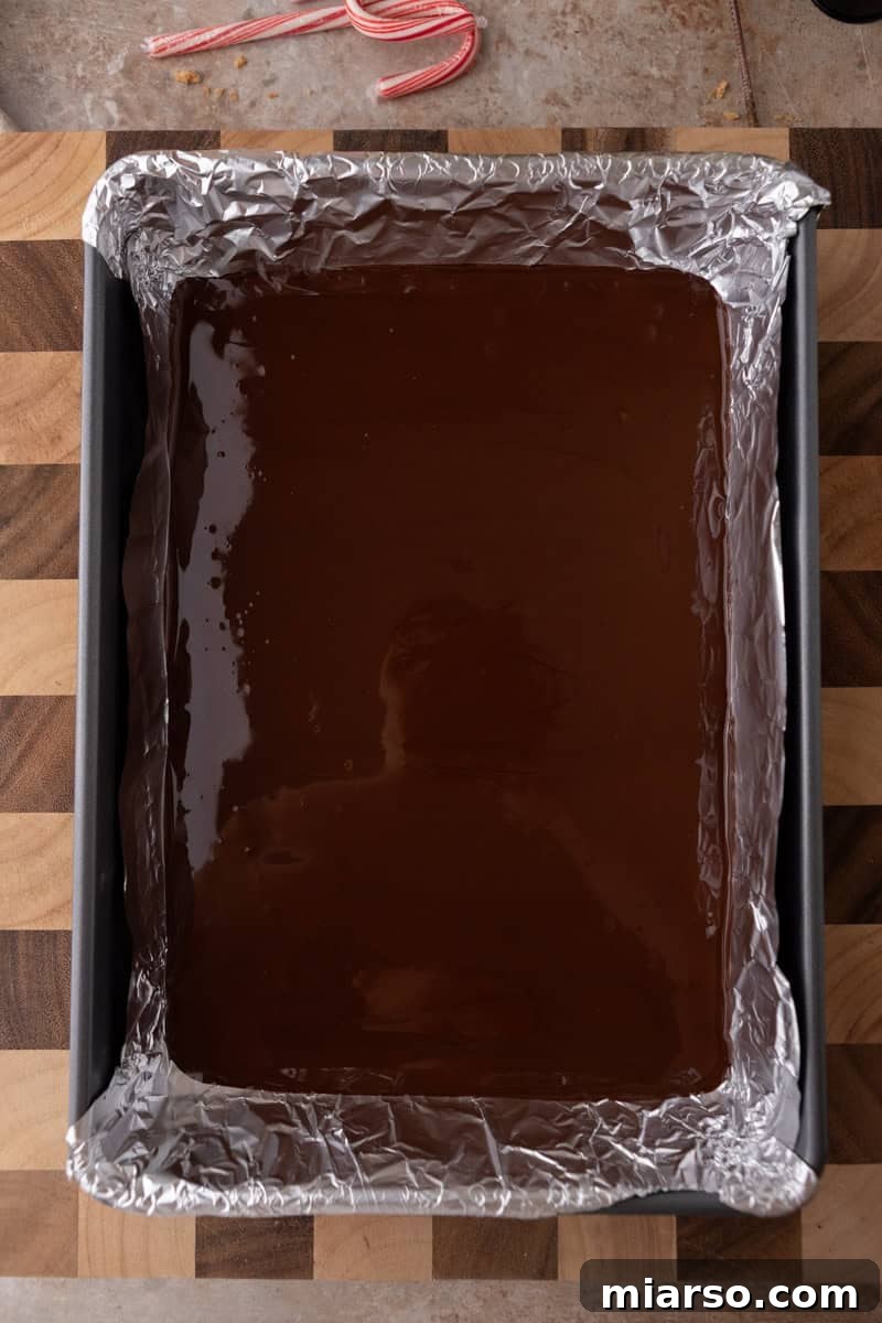  A baking pan lined with foil and filled with melted dark chocolate spread evenly, forming the base layer of the peppermint bark.