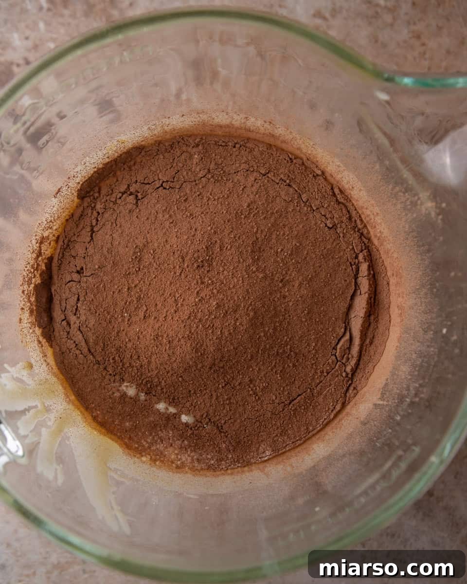 Ina Garten's Viral Brownie Pudding Sensation 6 Unsweetened cocoa powder and all-purpose flour being gently folded into the pale, fluffy egg and sugar batter in a large glass bowl.