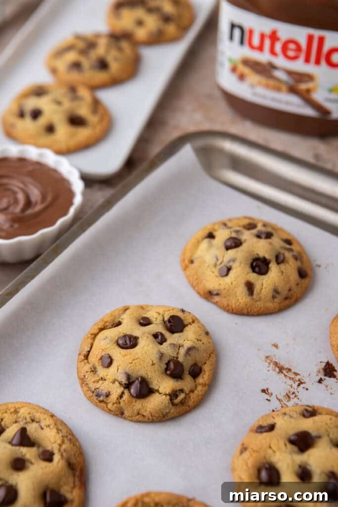Freshly baked Nutella-stuffed chocolate chip cookies cooling on a tray with Nutella jar in the background