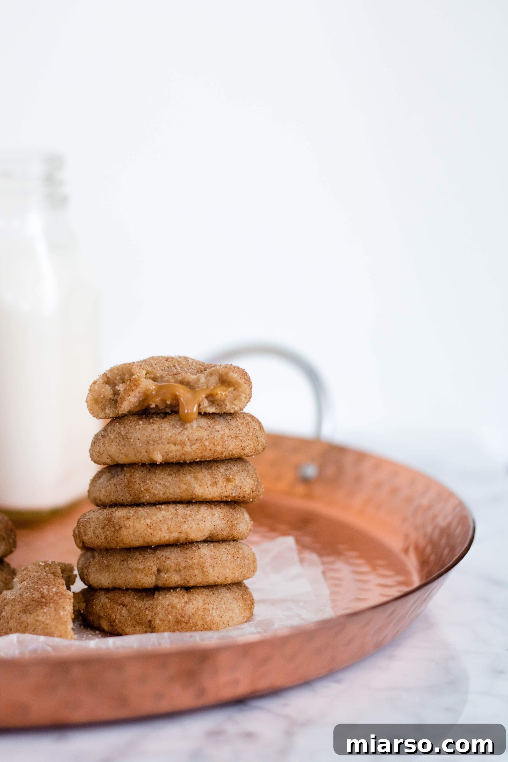 A freshly baked Snickerdoodle cookie with a light golden hue, covered in a beautiful cinnamon sugar crust. The edges are slightly crisp, while the center appears soft. A small amount of warm dulce de leche has gently seeped out, adding to its rustic charm. It rests on parchment paper on a baking sheet, with other cookies blurred in the background.