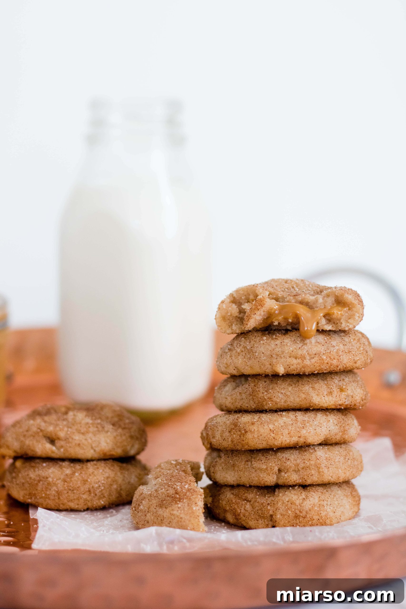 A stack of three golden Snickerdoodle cookies, generously coated in cinnamon sugar, with a peek of the creamy dulce de leche filling emerging from between them. The cookies are soft and inviting, positioned on a rustic wooden surface with a soft, warm light.