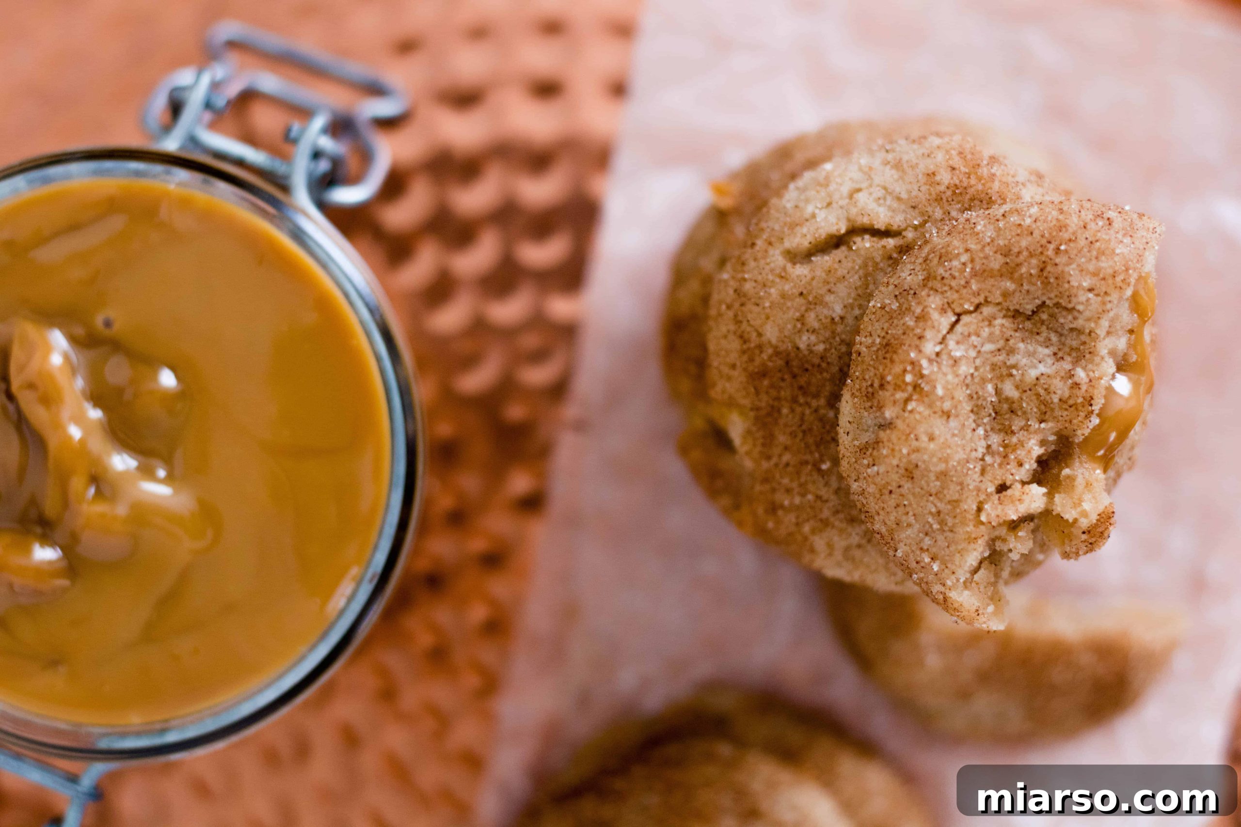 A wide shot of a batch of freshly baked Dulce de Leche Snickerdoodle cookies cooling on a wire rack, with some cookies showing the signature cinnamon sugar coating and others revealing the warm, caramel filling peeking out. The background is a rustic wooden surface, creating a cozy, inviting atmosphere.