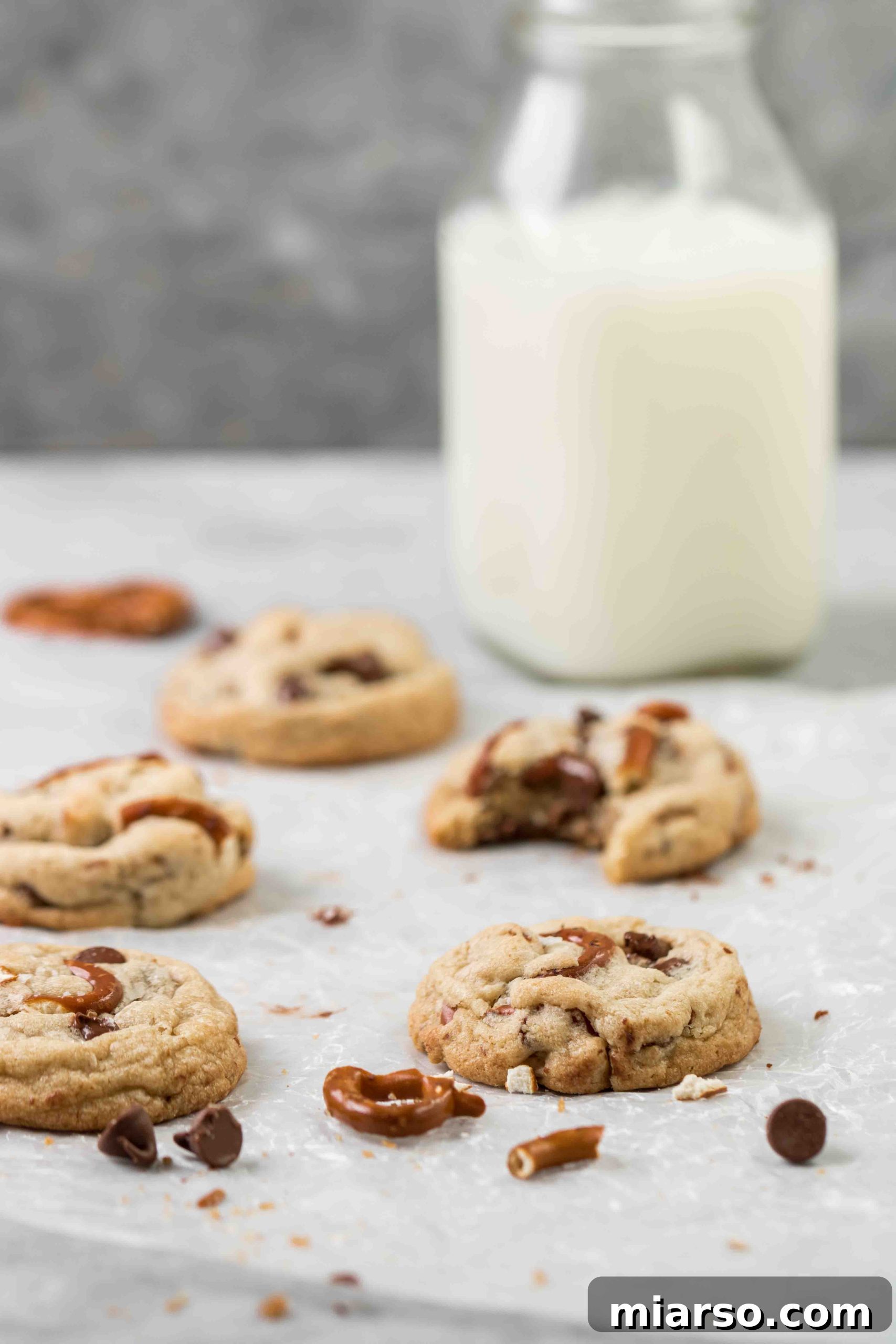 Three golden-brown pretzel chocolate chip cookies on a cooling rack next to a glass of milk, with visible pretzel pieces and melted chocolate.