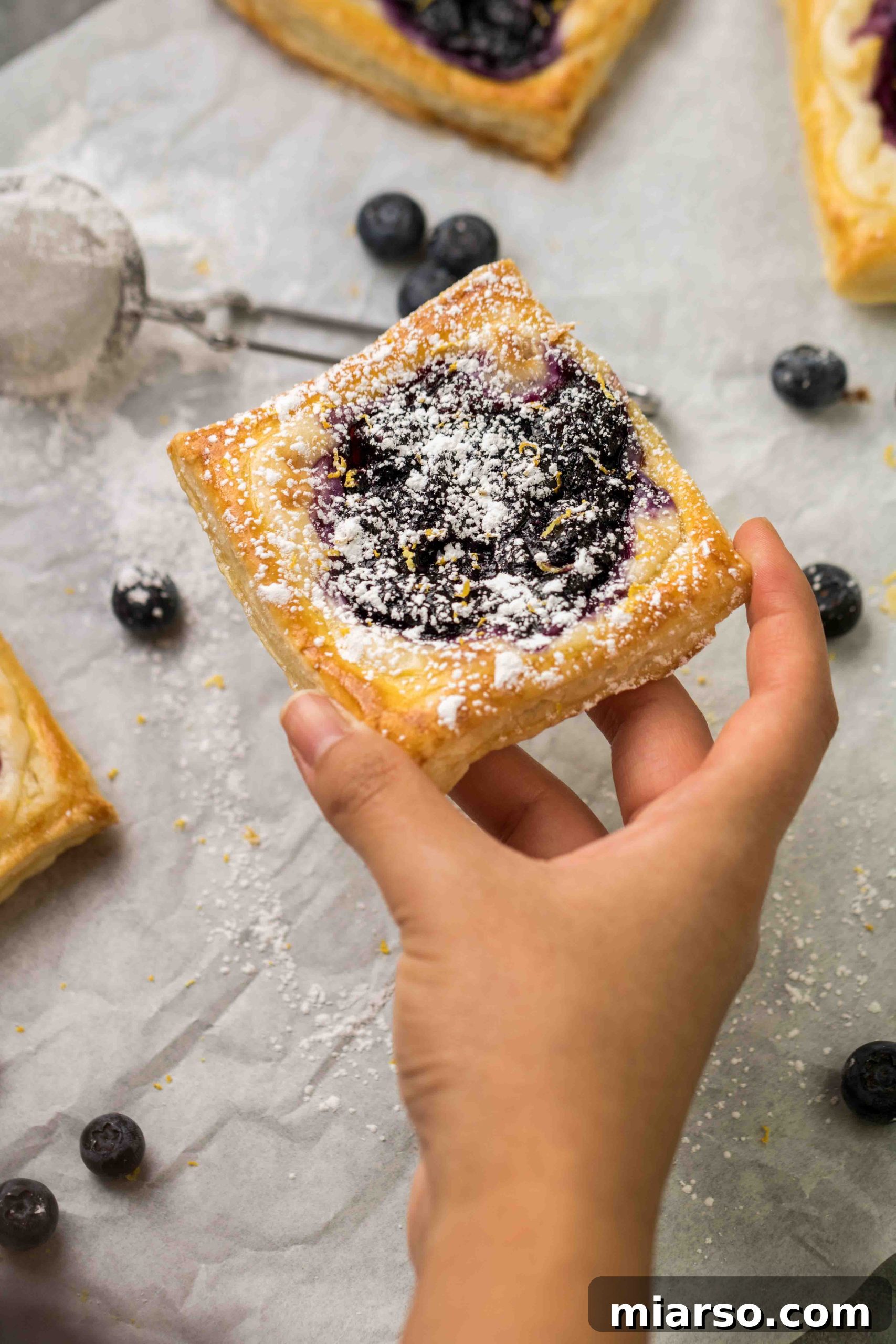 Flaky Blueberry Cheese Danish Made Easy 10 A hand holding a freshly baked blueberry cheese danish, showing its flaky texture and rich filling.