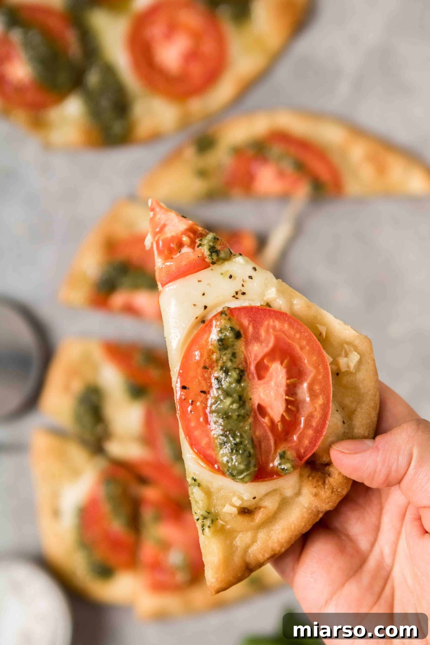 Hand holding a slice of golden naan bread pizza topped with fresh pesto