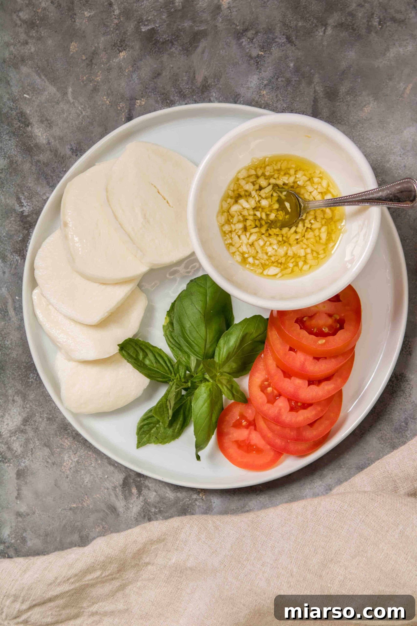 Fresh ingredients laid out for making pesto pizza: basil, mozzarella, tomato slices, and minced garlic in olive oil.