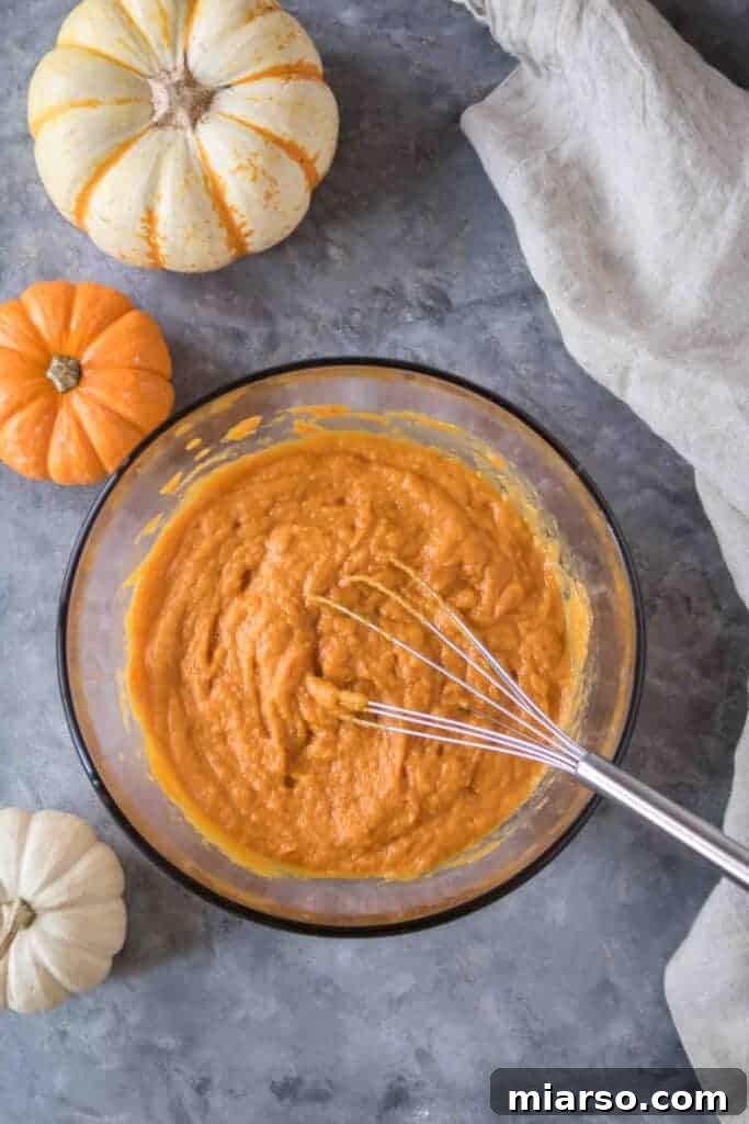 Pumpkin pie filling mixture whisked until smooth in a glass bowl surrounded by mini pumpkins on a gray surface.