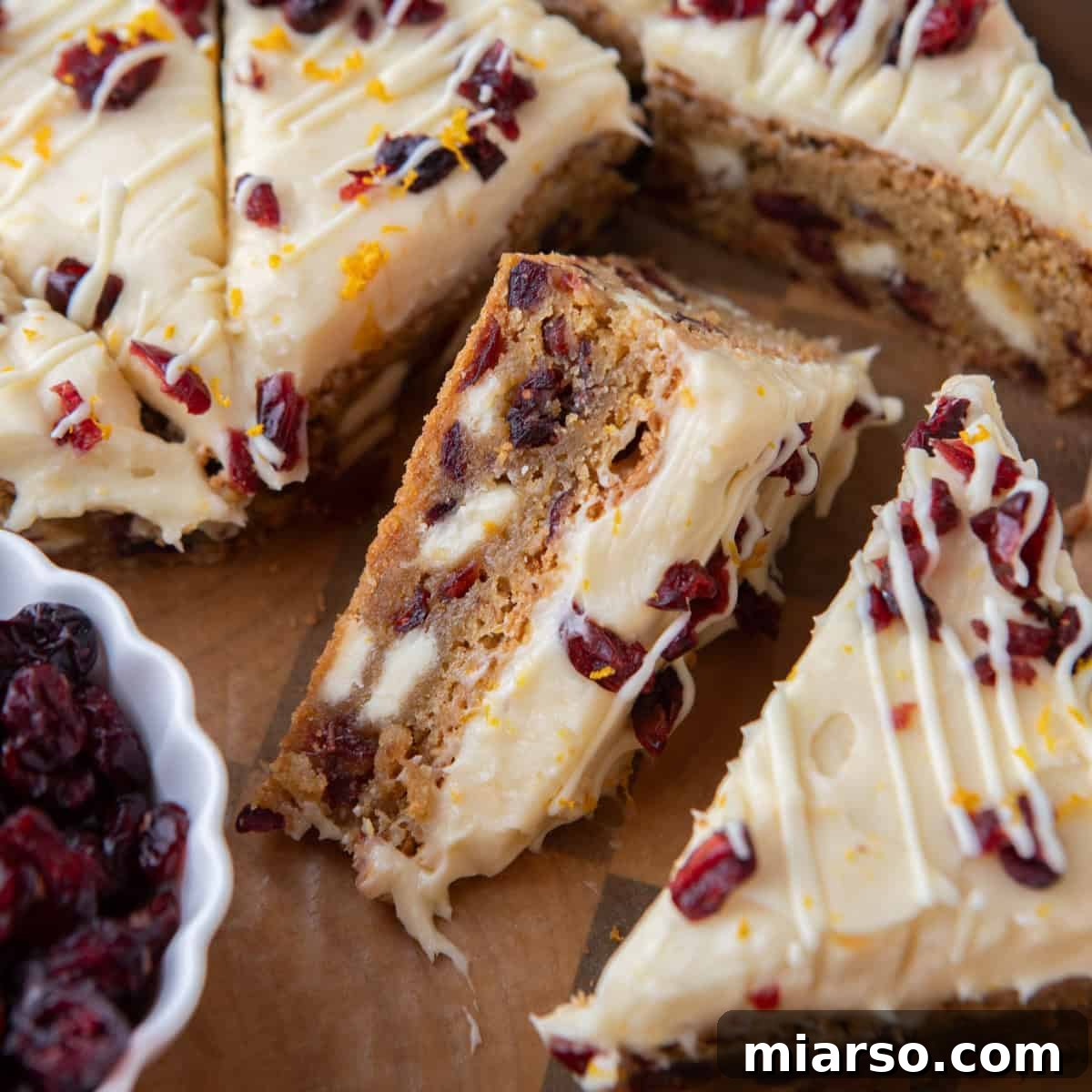 Close-up of homemade cranberry bliss bars topped with cream cheese frosting, dried cranberries, and orange zest drizzled with white chocolate.