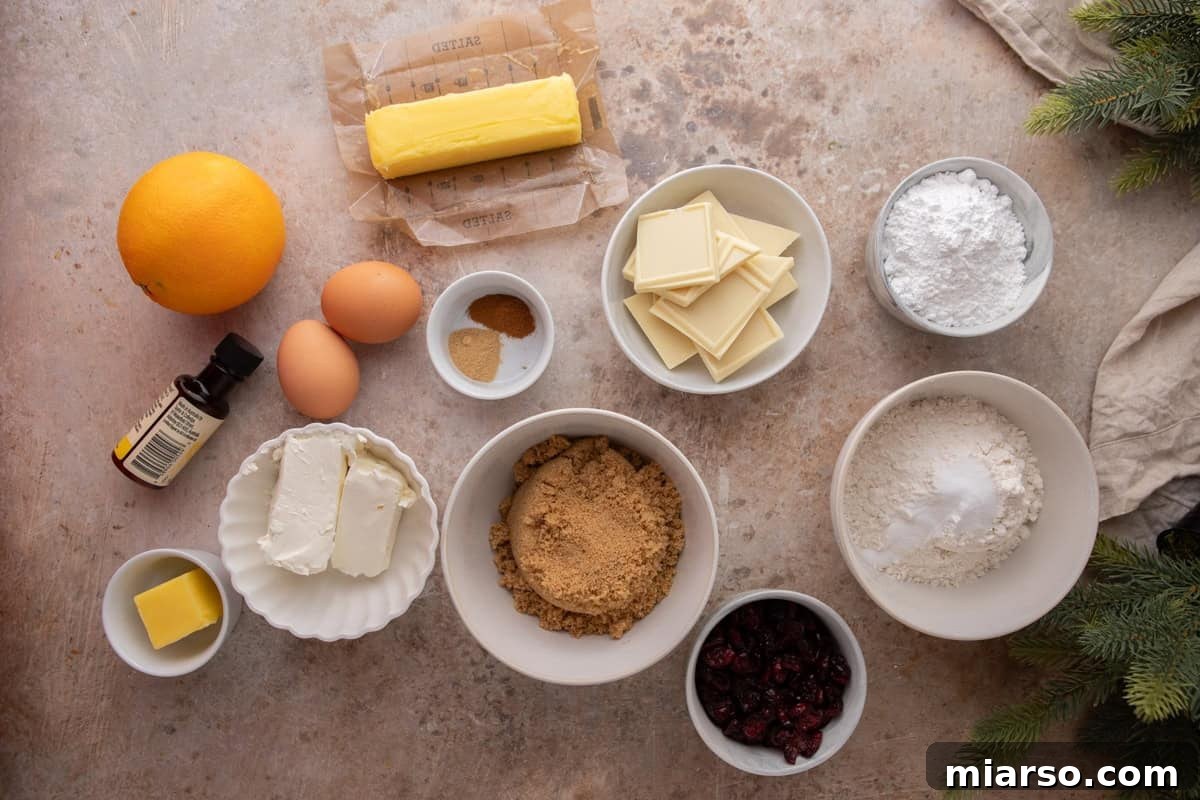 Overhead view of ingredients for cranberry bliss bars including butter, eggs, flour, brown sugar, dried cranberries, white chocolate, orange, spices, cream cheese, and powdered sugar arranged on a counter.

