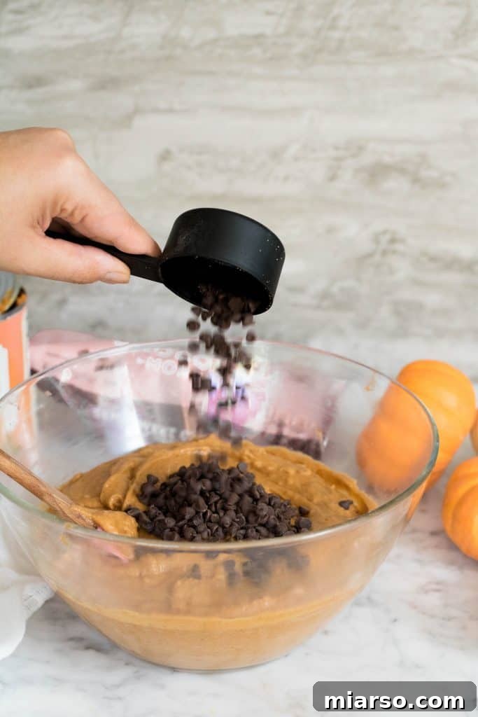 A generous handful of mini chocolate chips being dropped into the orange-hued pumpkin muffin batter, ready for a final gentle fold.