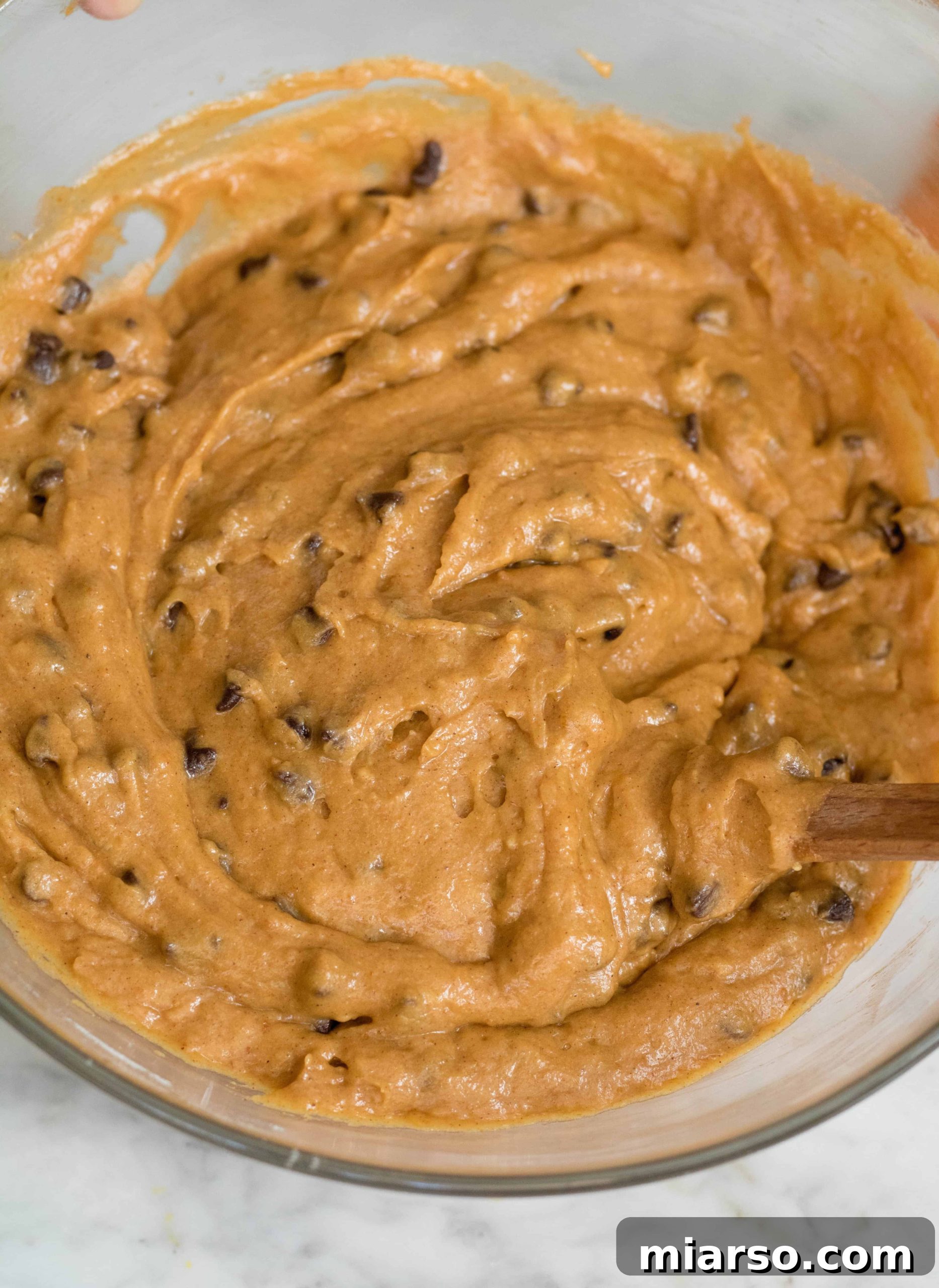 A bowl of freshly mixed orange pumpkin muffin batter, studded generously with mini chocolate chips, showcasing its perfect, slightly lumpy consistency.