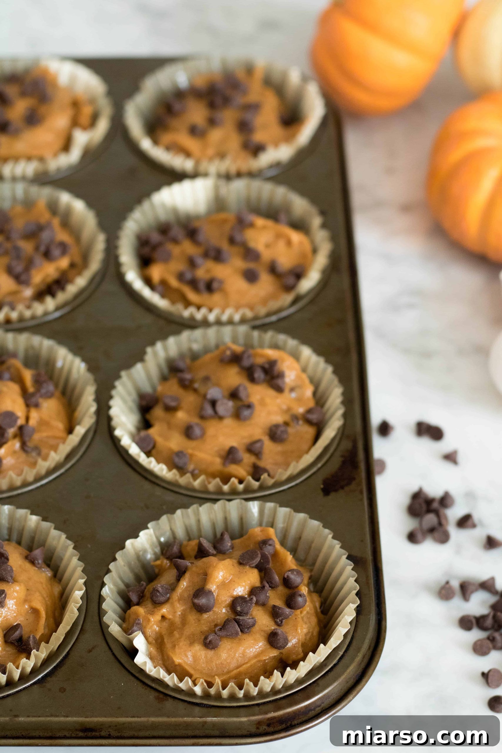 A muffin tin filled with raw pumpkin muffin batter in paper liners, dotted with chocolate chips and ready to be baked into delicious treats.