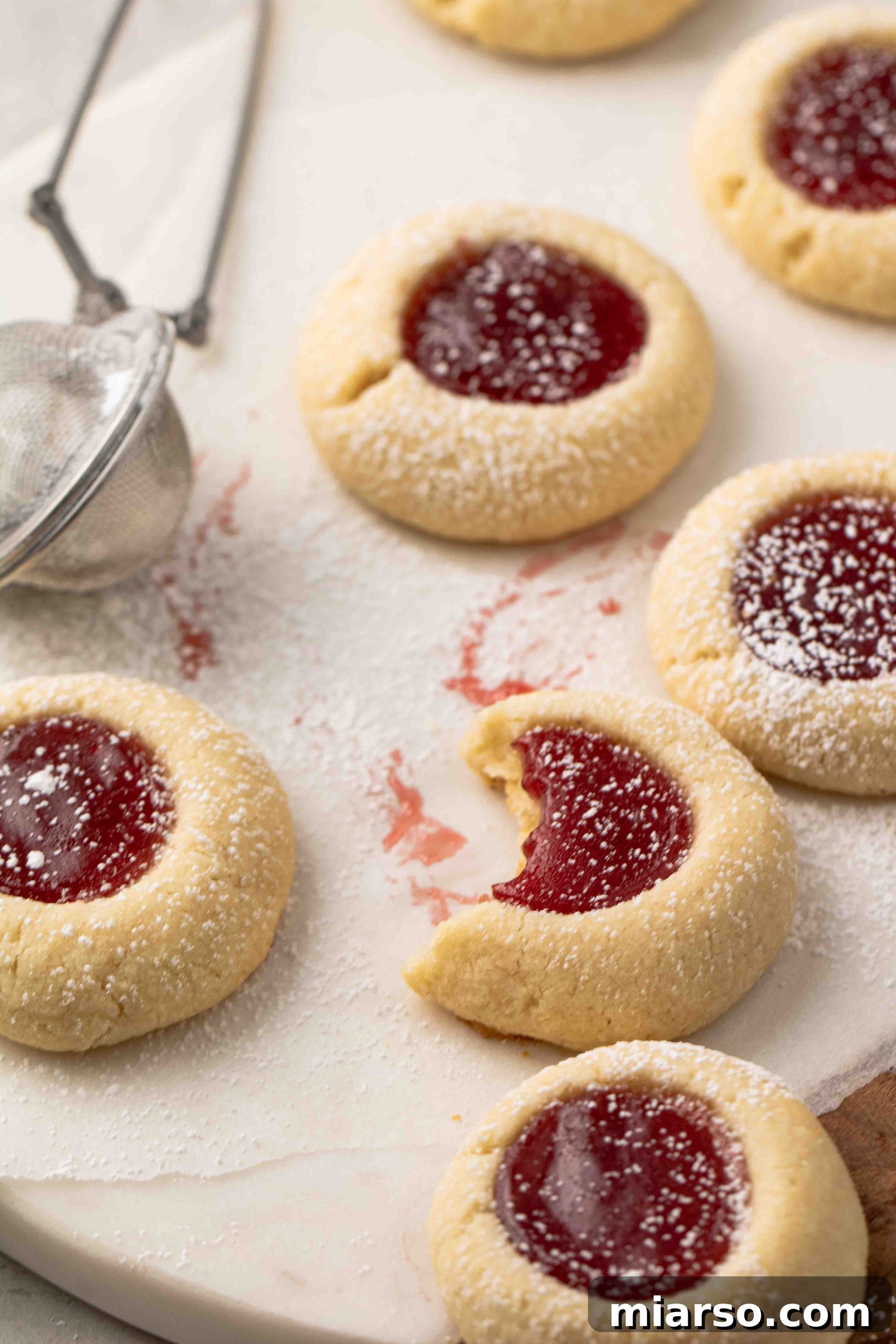Jam-Kissed Almond Thumbprint Cookies 13 Close-up shot of easy raspberry thumbprint cookies with glistening jam filling and a sprinkle of powdered sugar.