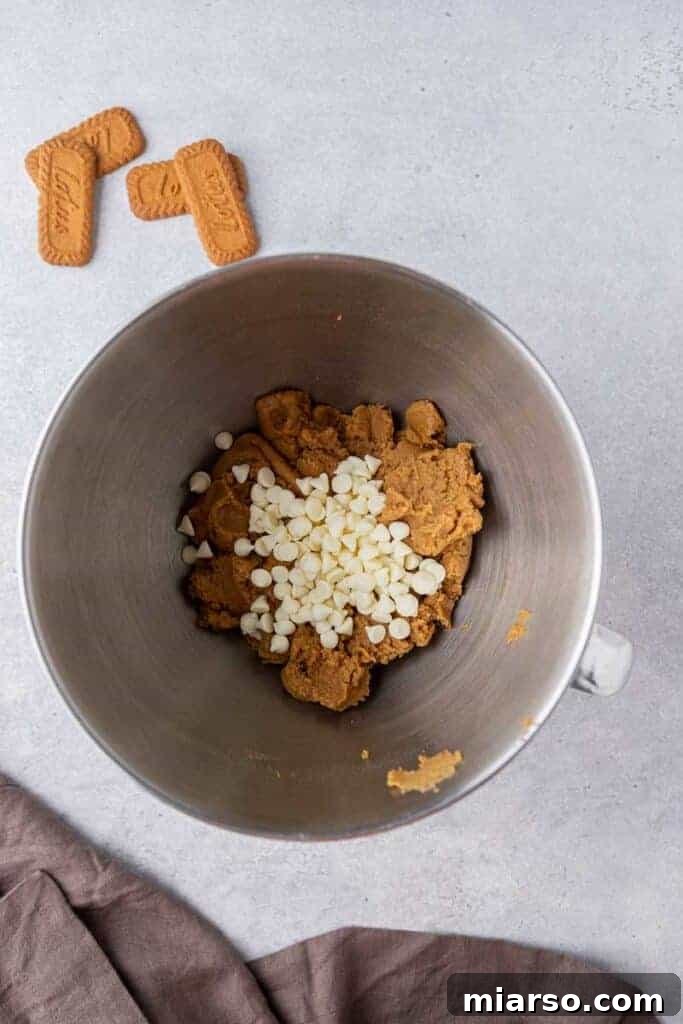 The Best Crumbl Biscoff White Chip Cookies Recipe 8 White chocolate chips being added to the cookie dough in a mixing bowl, ready to be folded in.