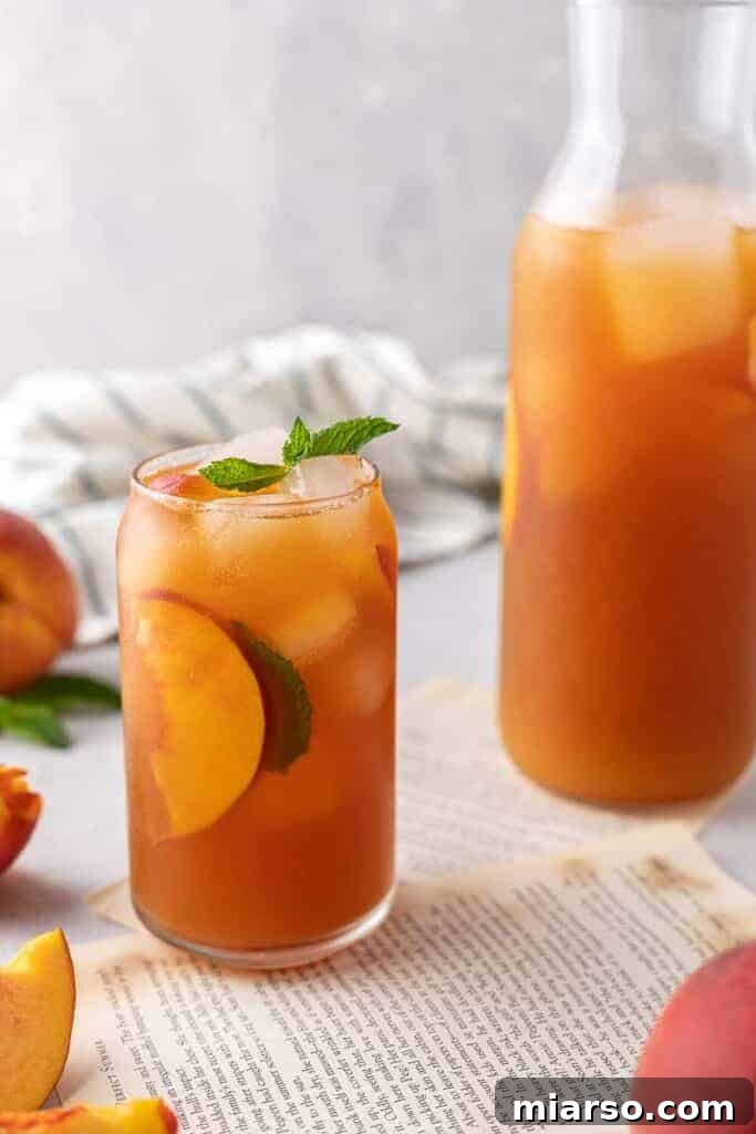 Ice cold homemade peach iced tea served in a glass next to a pitcher filled with more tea and fresh peach slices, ready for a refreshing summer day.