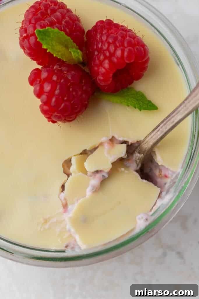A spoon cracking through the white chocolate layer on top of raspberry overnight oats.