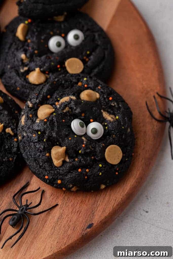 Close-up shot of several black Halloween monster cookies with colorful sprinkles and edible eyeballs, stacked on a cooling rack.