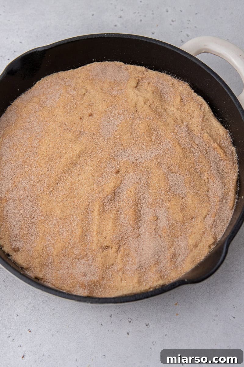 Snickerdoodle cookie dough pressed into a cast iron skillet, sprinkled with cinnamon sugar, ready for baking.