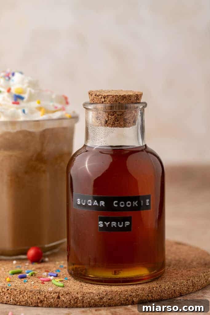 A clear glass container filled with homemade Starbucks Sugar Cookie Syrup, garnished with a sprig of evergreen and a few festive sugar sprinkles, set against a cozy, blurred holiday background.