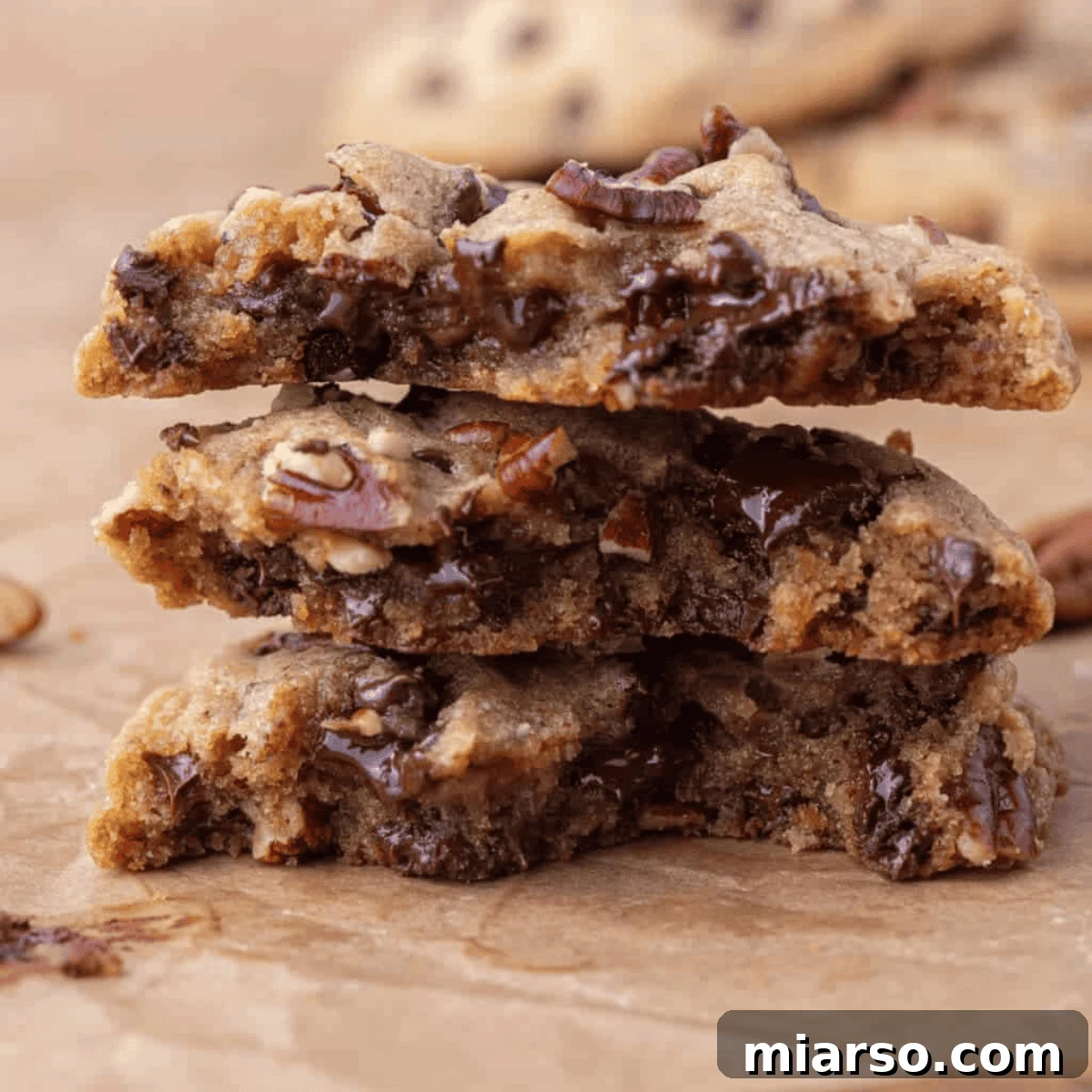 Stack of brown butter pecan chocolate chip cookies broken in half, showing gooey melted chocolate chips and toasted pecan pieces on parchment paper.
