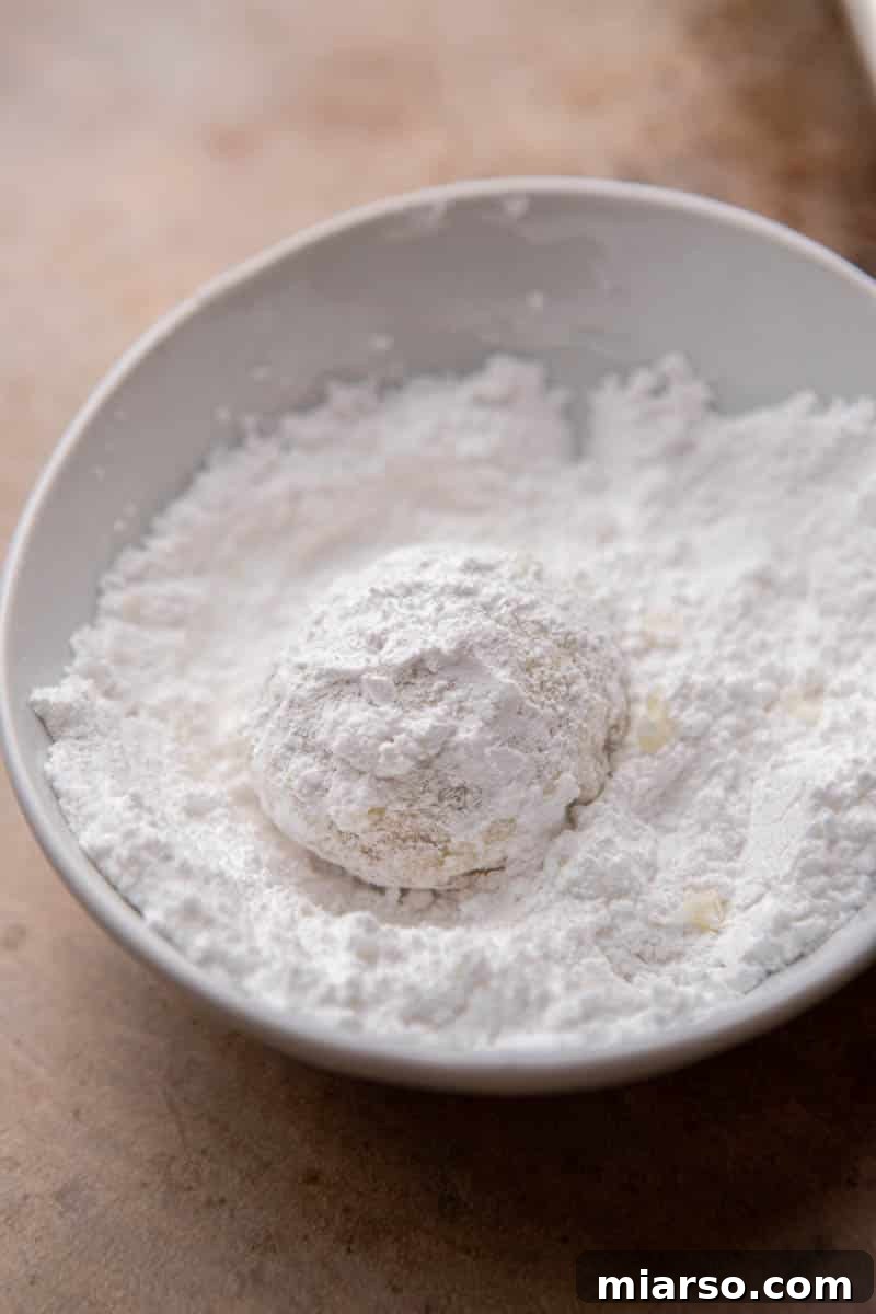 Melt-in-Your-Mouth Snowball Perfection 10 A warm cookie being dipped into a bowl of powdered sugar for its first coating, demonstrating the traditional finishing touch.