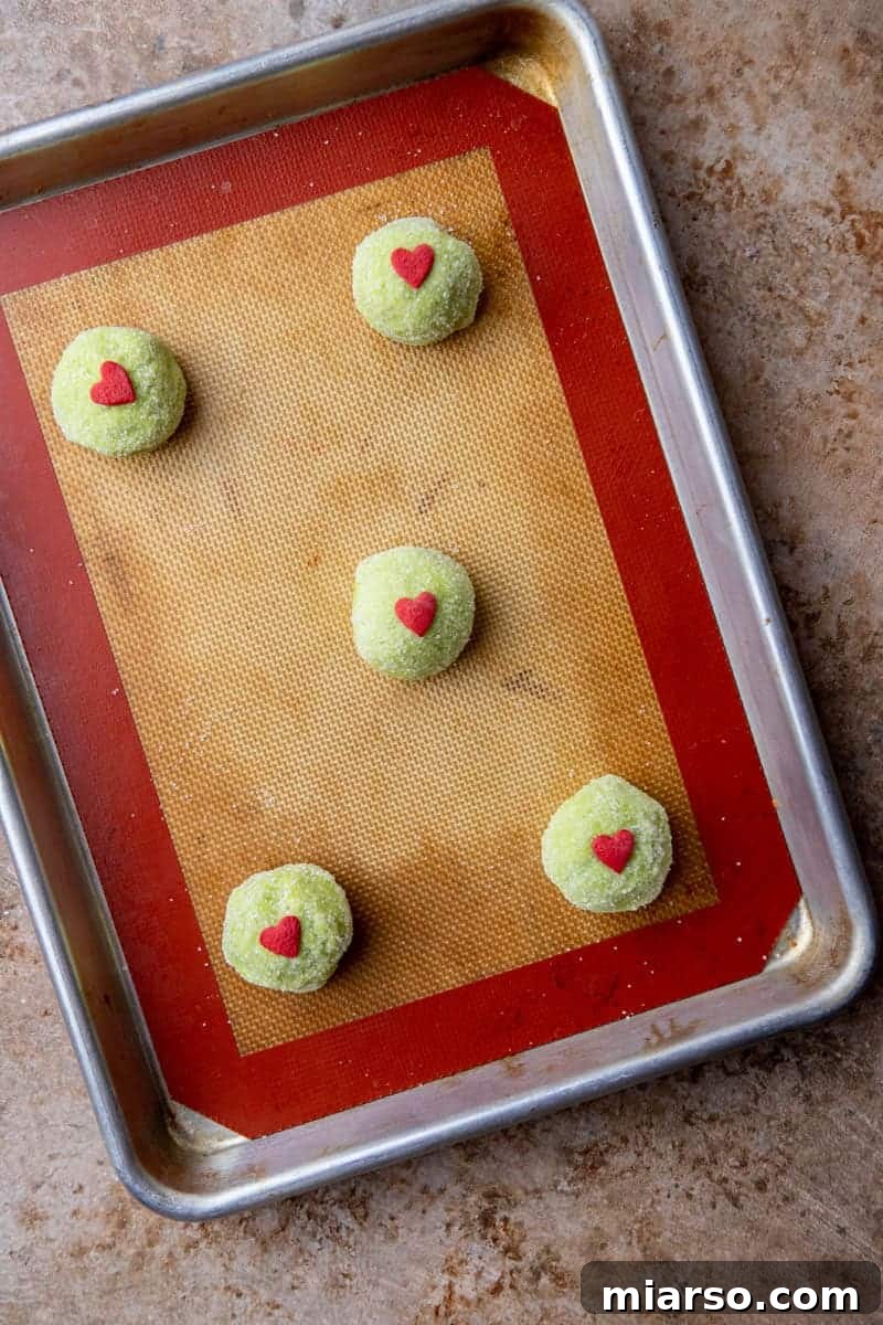 Grinch's Heart-Melting Sugar Cookies 9 Raw cookie dough balls with heart sprinkles on a baking sheet, pre-oven