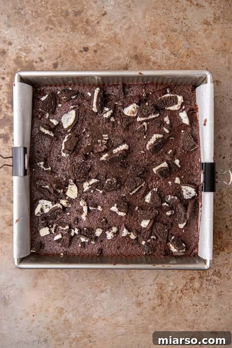 Decadent Oreo Fudge Brownies 9 Unbaked brownie batter layered with Oreos and topped with crushed cookies, in a parchment-lined pan.