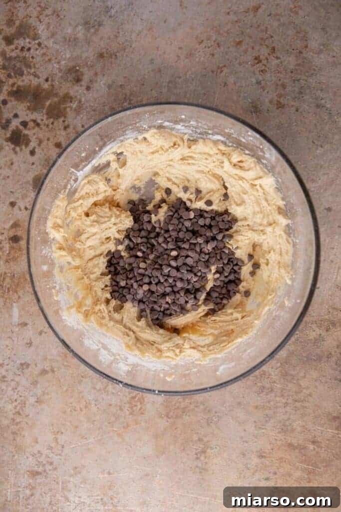 Mini chocolate chips being added to soft sugar cookie dough in a mixing bowl, ready to be incorporated.