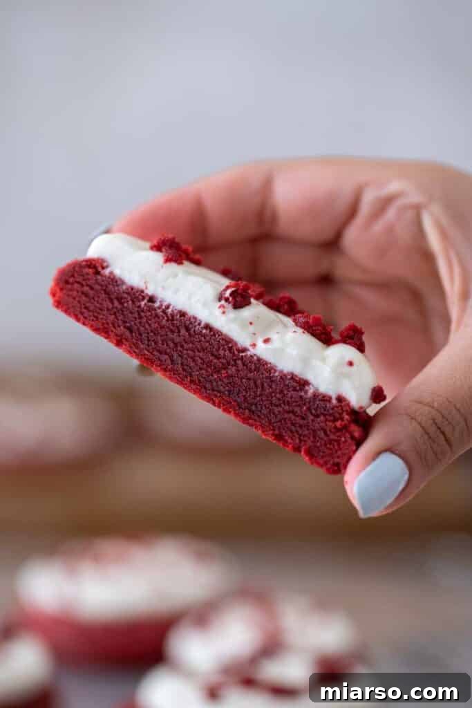 A hand holding a Crumbl red velvet cookie that has been sliced in half, showcasing its thick, moist texture and generous frosting layer.
