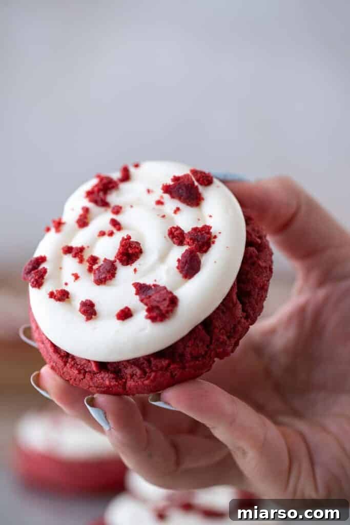 A hand holding a perfectly baked Crumbl red velvet cupcake cookie with a generous swirl of cream cheese frosting.