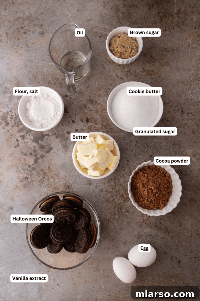 A collection of brownie ingredients laid out on a kitchen counter, including butter, oil, sugars, cocoa powder, eggs, vanilla, flour, and a pack of Halloween Oreos.