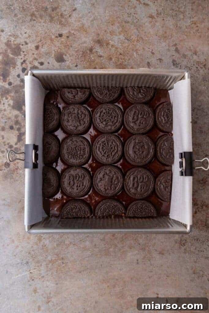 A layer of whole and halved Halloween Oreos arranged on top of the first layer of brownie batter in a baking pan.