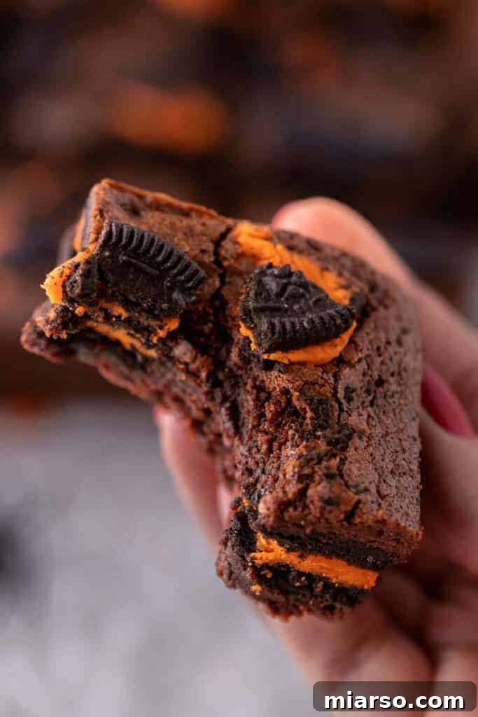 A hand holding a perfectly sliced, fudgy Halloween Oreo brownie, showcasing the rich chocolate and orange Oreo filling.
