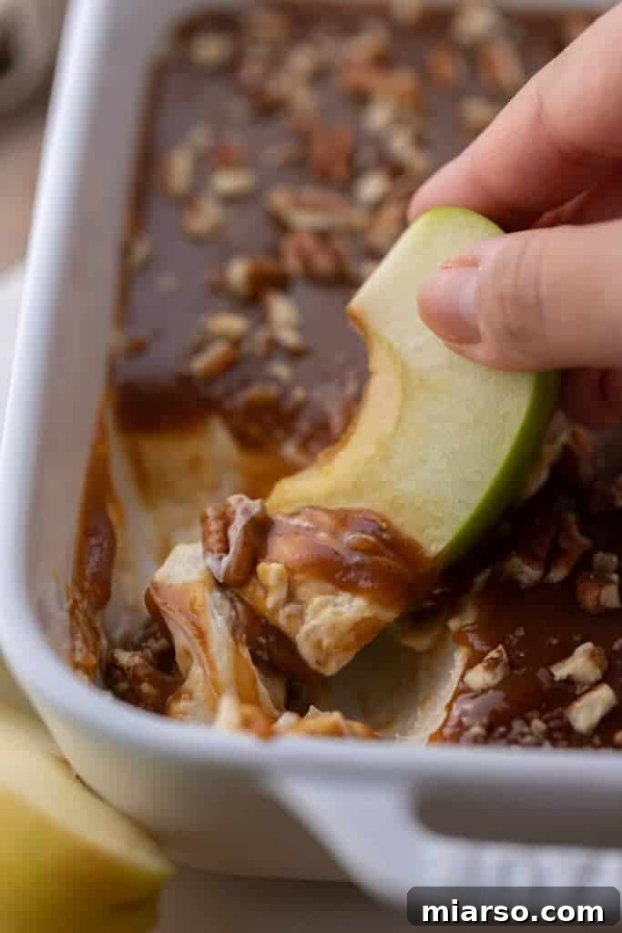 A hand dipping a fresh apple slice into the creamy caramel apple cheesecake dip.