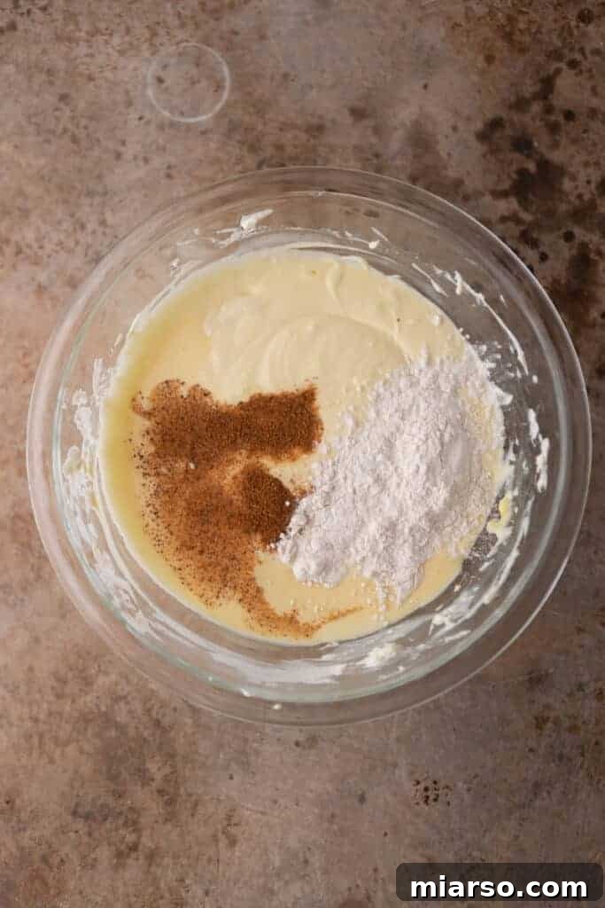 Dry ingredients and spices on top of cheesecake batter in a bowl, ready to be mixed