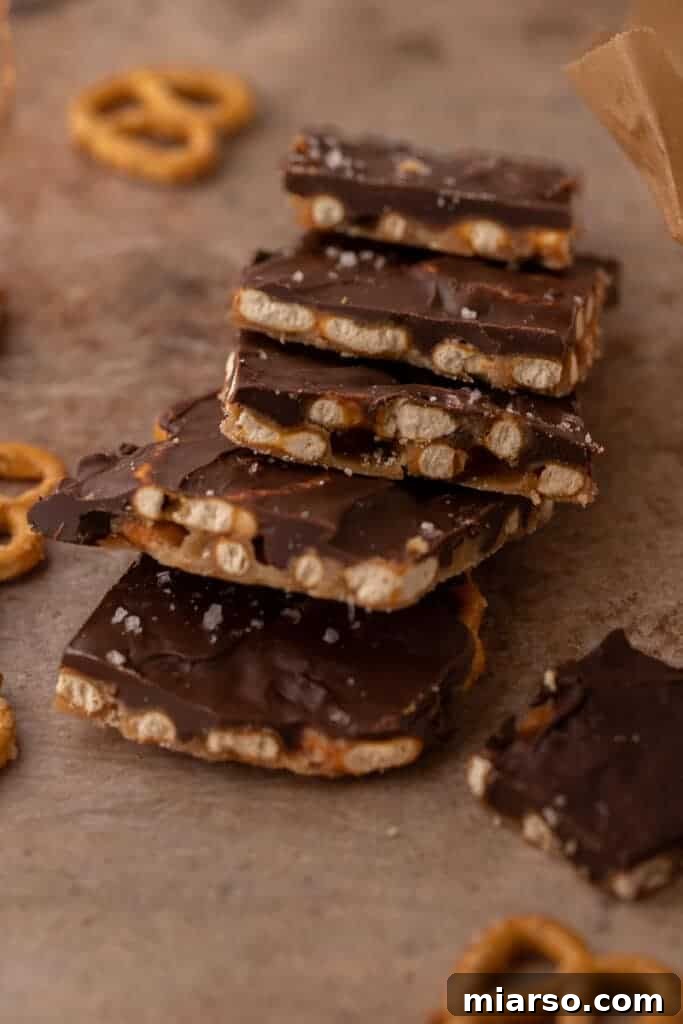 A close-up view of a stacked pile of Chocolate Caramel Pretzel Bark pieces, glistening with smooth chocolate and a delicate sprinkle of flaky sea salt.