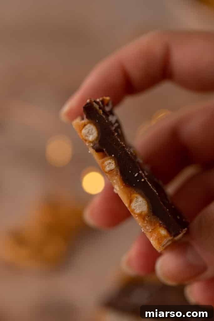 A hand holds a tempting piece of Christmas Chocolate Pretzel Treat, clearly showing the distinct layers of crunchy pretzel and gooey caramel, all enrobed in chocolate.
