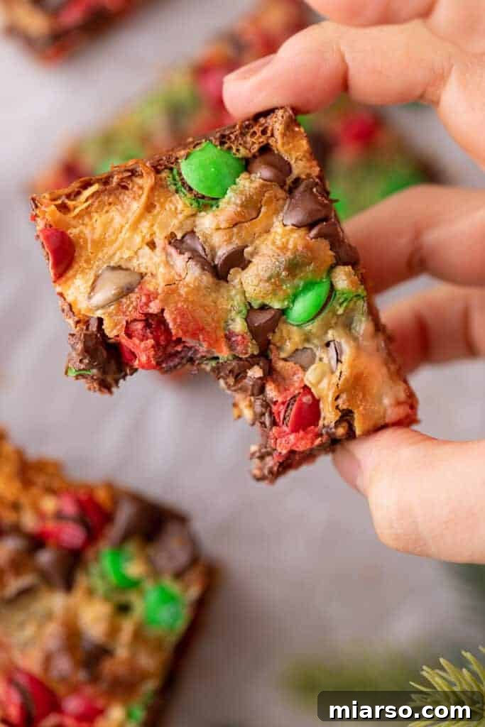 A hand holding up a Christmas magic cookie bar with a bite taken out, showing the layers inside