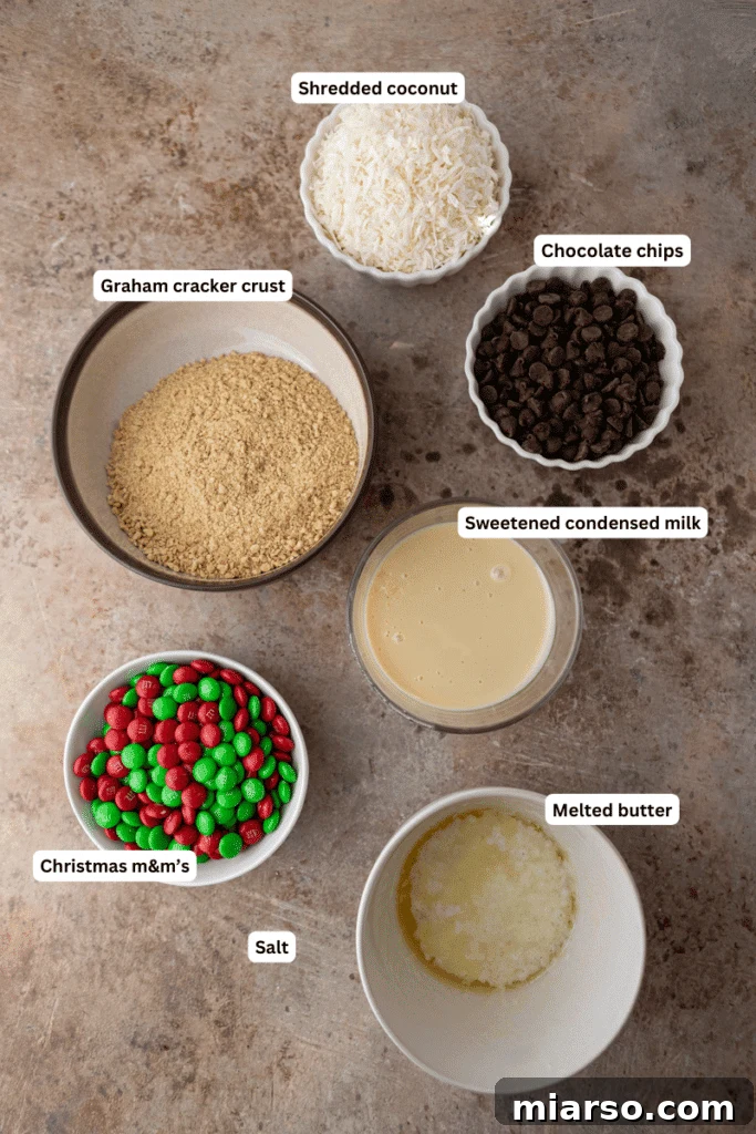 Ingredients for making the Christmas magic cookie bars laid out on a surface, including butter, graham crackers, chocolate chips, and M&Ms