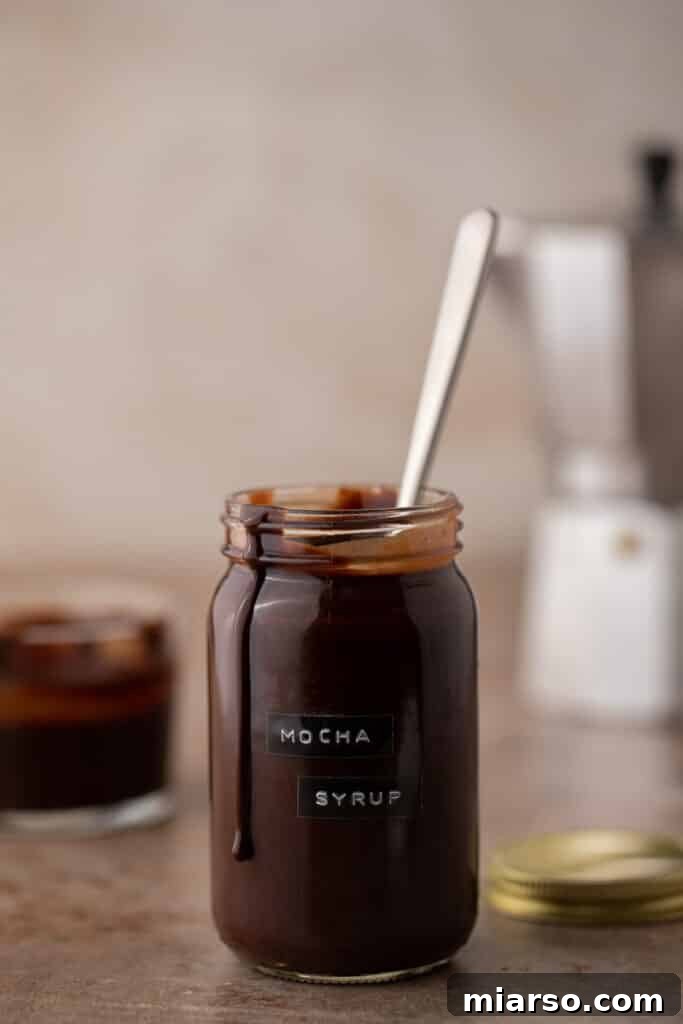 Ultimate Homemade Mocha Coffee Syrup 9 A beautifully composed shot of homemade mocha coffee syrup in a jar with coffee beans and chocolate pieces.