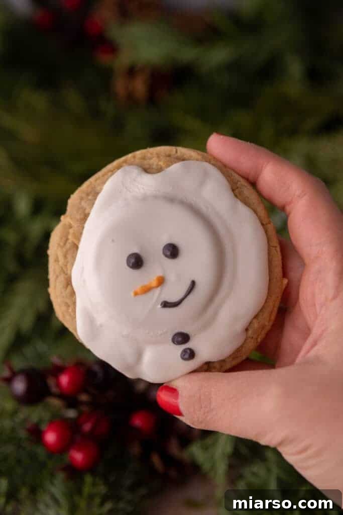 A hand holding up a single, beautifully decorated melted snowman peanut butter cookie, showcasing its festive detail.