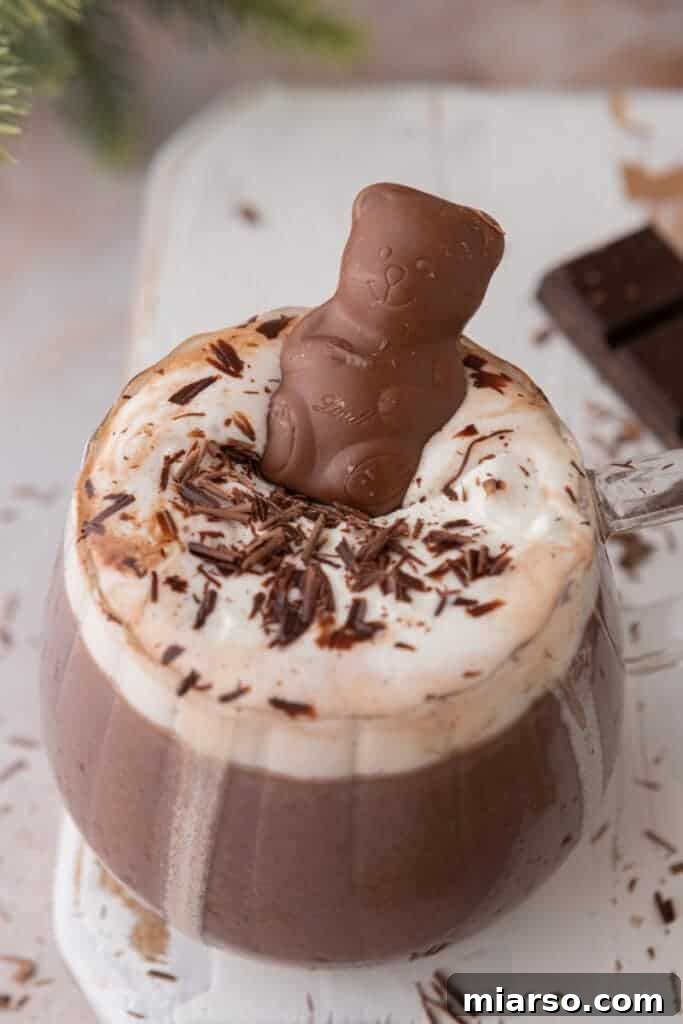 A cute chocolate bear floating on whipped cream in a glass mug of hot chocolate, with chocolate shavings.