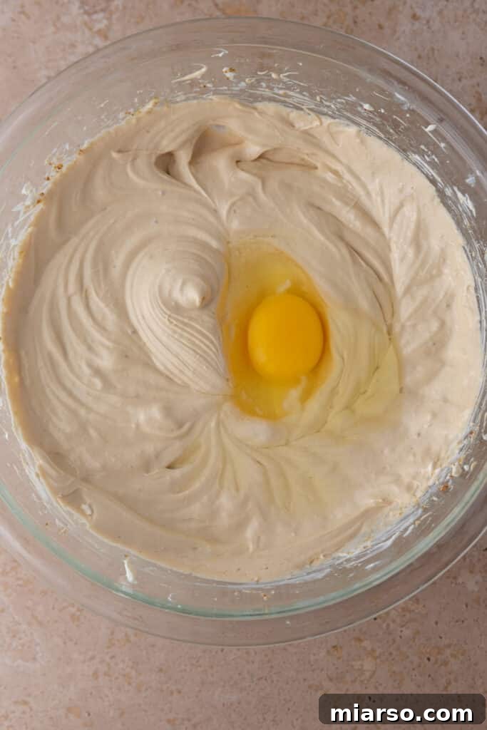 A cracked egg sitting atop the smooth gingerbread cheesecake batter in a mixing bowl.