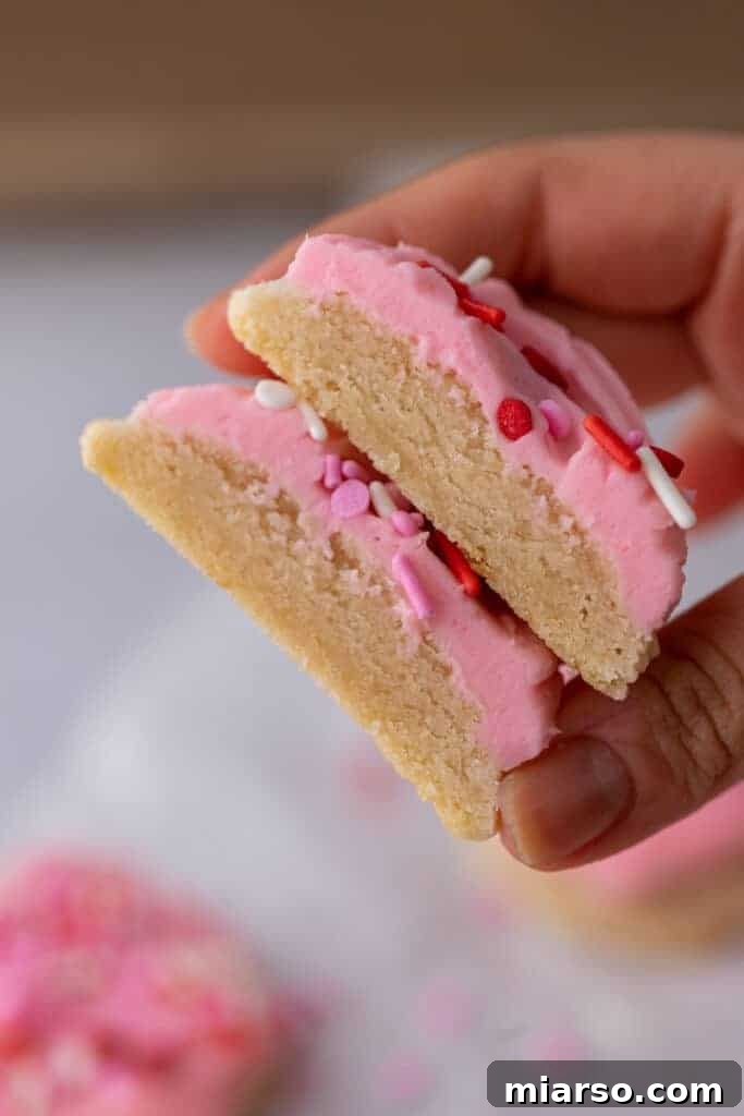 Ultimate Valentine Frosted Sugar Cookies 11 A hand holding a Valentine's Day frosted sugar cookie cut in half, showing its soft texture