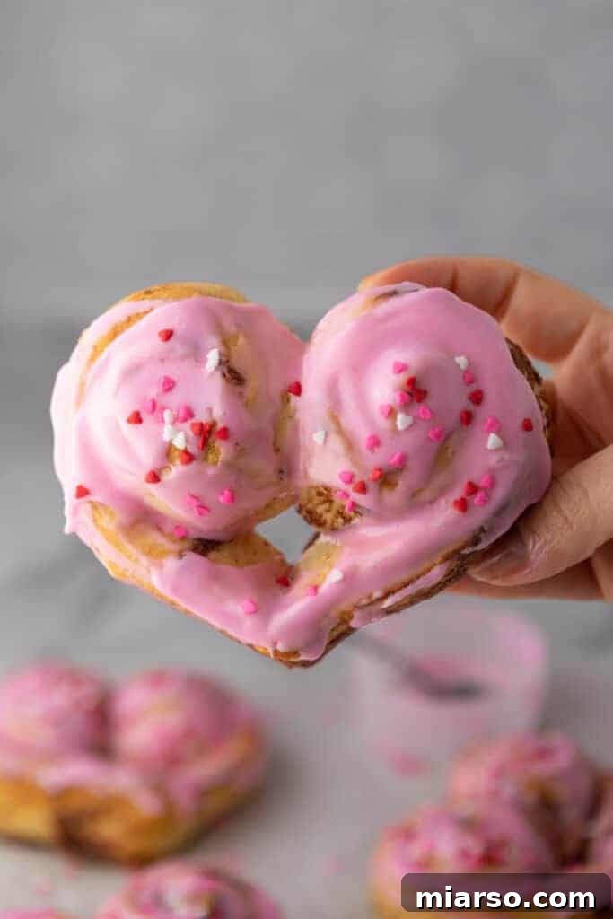 Valentines Sweetheart Cinnamon Buns Made Easy 2 A Valentine's Day heart-shaped cinnamon roll, held delicately by a hand, showcasing its vibrant pink frosting and sprinkles, perfect for a romantic breakfast.